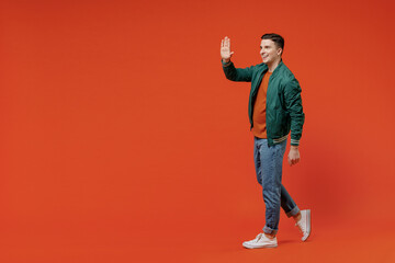 Fototapeta na wymiar Full size body length side view profile young brunet man 20s wears red t-shirt green jacket go move meet greet waving hand isolated on plain orange background studio portrait. People emotions concept.