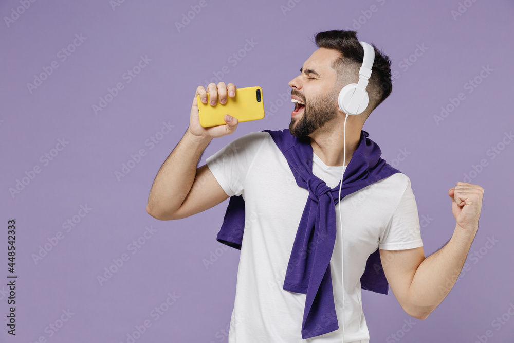 Wall mural Side view profile young brunet man 20s wears white t-shirt purple shirt listen to music in headphones keep eyes closed sing in mobile cell phone isolated on pastel violet background studio portrait