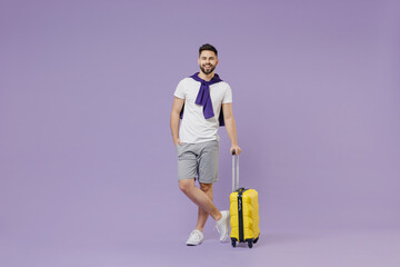 Full size body length side view fun traveler tourist young brunet man 20s wear white t-shirt purple shirt hold yellow suitcase bag hand in pocket isolated on pastel violet background studio portrait.