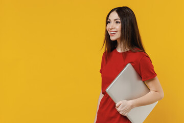 Smiling excited vivid charming fun happy cheerful young brunette woman 20s wears basic red t-shirt hold under hand laptop pc computer looking ahead aside isolated on yellow background studio portrait.