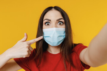 Close up young brunette woman 20s wear basic red t-shirt point on sterile face mask ppe to safe from coronavirus covid-19 flu do selfie shot on mobile phone isolated yellow background studio portrait