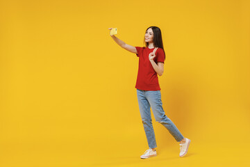Fototapeta na wymiar Full size body length young brunette woman 20s wears basic red t-shirt do selfie shot on mobile cell phone post photo on social network show victory sign isolated on yellow background studio portrait