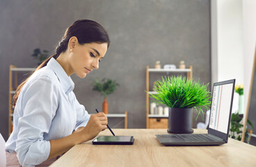 Elegant young business woman working with modern digital tablet and laptop in her office. Side view of a serious woman sitting at a table and checking documents on a tablet using a stylus pen.