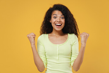 Overjoyed african american young woman 20s wears green shirt doing winner gesture celebrating clenching fists say yes isolated on yellow background studio portrait. People emotions lifestyle concept