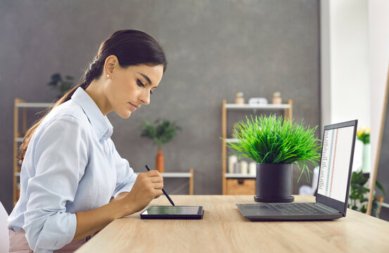 Elegant young business woman working with modern digital tablet and laptop in her office. Side view of a serious woman sitting at a table and checking documents on a tablet using a stylus pen.