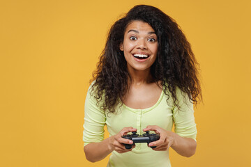 Gambling smiling fun african american young woman 20s wear green shirt hold in hand play pc game with joystick console isolated on yellow background studio portrait. People emotions lifestyle concept © ViDi Studio