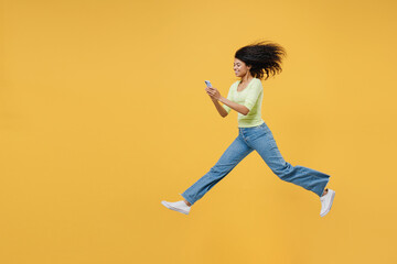 Full size body length side view overjoyed happy african american young woman 20 wear green shirt jump hold in hand use mobile cell phone keep eyes closed isolated on yellow background studio portrait