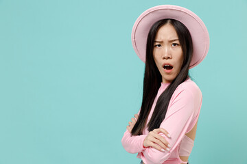 Displeased sad young brunette asian woman 20s with open mouth wear pink clothes hat hold hands crossed stand sideways isolated on pastel blue color background studio portrait. People emotions concept