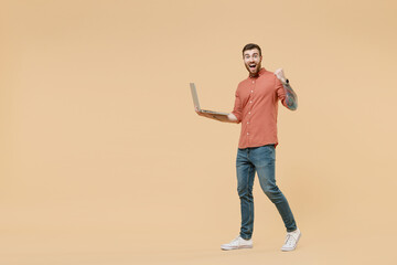 Full size body length fun tatooed young brunet man 20s short haircut wears apricot shirt go move hold use laptop pc computer clench fists say yes isolated on pastel orange background studio portrait