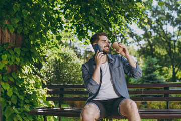 Young happy man in blue shirt sit on bench talk by mobile cell phone look aside hold takeaway craft...
