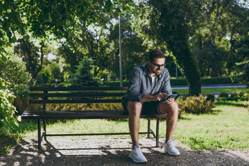 Full length young smiling man 20s in blue shirt glasses sit on bench using mobile cell phone chat rest relax in spring green city park sunshine lawn outdoors on nature Urban lifestyle leisure concept.