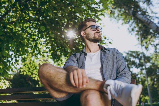 Bottom View Young Calm Smiling Happy Man 20s In Blue Shirt Shorts Glasses Sitting On Wooden Bench Look Overhead Rest Relax In Spring Green City Park Outdoors On Nature Urban Lifestyle Leisure Concept.