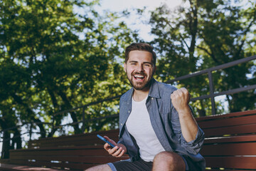 Young overjoyed man in blue shirt shorts sit on bench use mobile phone do winner gesture clench fist rest relax in spring green city sunshine park outdoor on nature Urban leisure lifestyle concept.