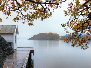 White vintage cabin  by the sea coast with wet pier . Foliage gold colors. Rainy , foggy day. Blurred background. Southern Norway