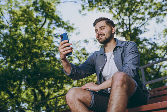 Bottom view young smiling happy man 20s in blue shirt shorts sit on bench do selfie shot on mobile cell phone rest relax in spring green city park outdoor on nature Urban leisure lifestyle concept