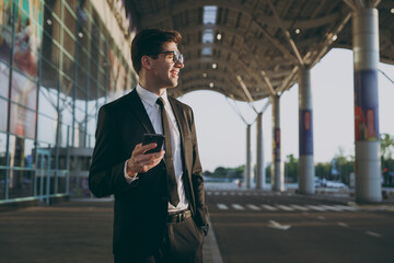 Bottom view traveler businessman young man wear black suit glasses stand outside at international airport terminal use mobile phone book taxi order hotel look aside Air flight business trip concept.