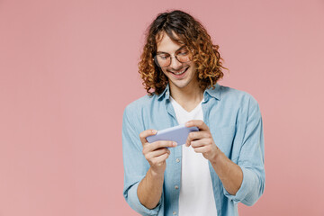 Young gambling man 20s with long curly hair in blue shirt glasses using play racing app on mobile cell phone hold gadget smartphone for pc video games isolated on pastel plain pink background studio