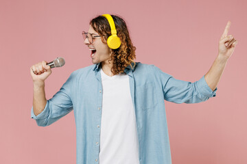 Young singer expressive man 20s with long curly hair wearing blue shirt white t-shirt glasses headphones sing song in microphone isolated on pastel plain pink color wall background studio portrait