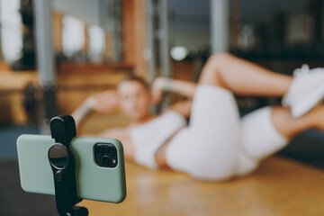 Full body blur young sporty woman in white sportswear warm up train do abdominal abs exercises at floor pump press with online instructor in gym indoors. Workout sport concept. Focus on mobile phone.