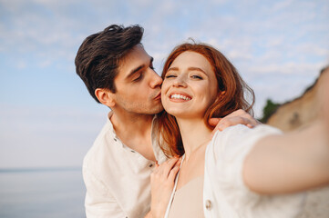 Close up happy tender young couple two family man woman in casual clothes hug each other do selfie shot pov on mobile phone kiss at sunrise over sea beach outdoor seaside in summer day sunset evening.