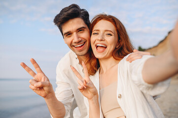Obraz premium Close up young couple two family man woman in white clothes hug each other do selfie shot pov on mobile phone show v-sign gesture at sunrise over sea beach outside seaside in summer day sunset evening