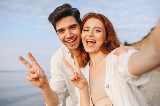 Close up young couple two family man woman in white clothes hug each other do selfie shot pov on mobile phone show v-sign gesture at sunrise over sea beach outside seaside in summer day sunset evening