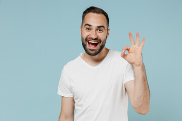 Young smiling happy satisfied man 20s in blank print design casual white t-shirt show ok okay gesture isolated on plain pastel light blue color background studio portrait. People lifestyle concept