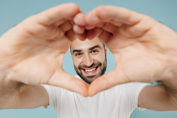 Close up young smiling romantic happy fun man 20s in casual white t-shirt showing shape heart look through hands heart-shape sign isolated on plain pastel light blue color background studio portrait