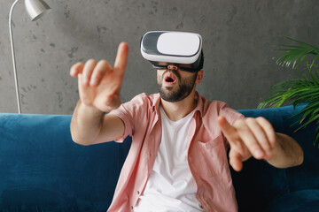 Naklejka premium Young smiling happy man wear casual clothes watching in vr headset pc gadget press button sitting on blue sofa at home flat indoors rest relax on weekends free time. People lounge lifestyle concept