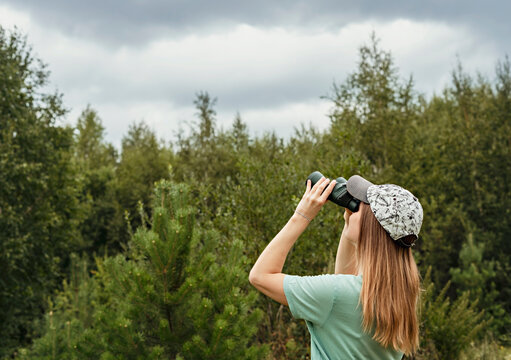 Young blonde woman bird watcher in cap and blue looking through binoculars at cloudy sky in summer forest ornithological research Birdwatching, zoology, nature, observation of animals