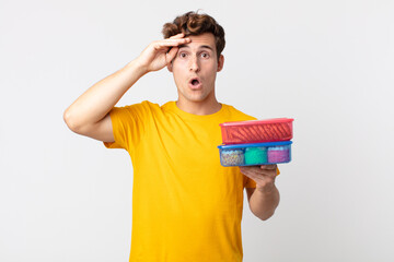 young handsome man looking happy, astonished and surprised and holding lunch boxes
