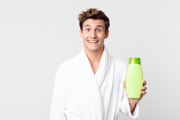 young handsome man looking happy and pleasantly surprised with bathrobe and holding a shampoo bottle - obrazy, fototapety, plakaty