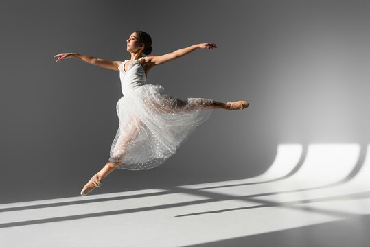 Side view of ballerina in white dress jumping in sunlight on grey background