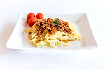 Spaghetti bolognese served on a white plate on a white background with tomatoes and basil.
