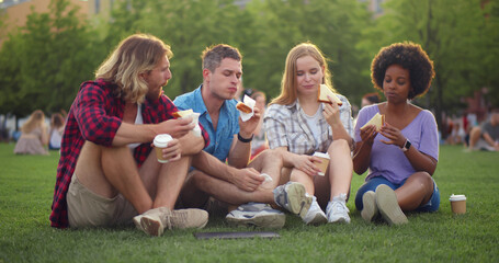 Fototapeta premium Diverse group of young people sitting on grass and eating sandwiches with coffee during picnic in park