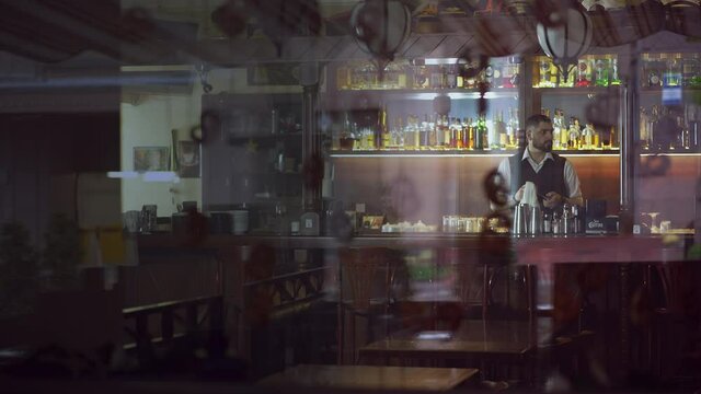 Bartender at the bar is wiping a glass. Wide shot through the glass at the bartender at work, waiting for visitors.
