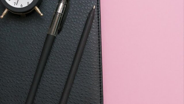 Black notepad with a pen and an alarm clock on a pink background. Use of wallpaper for education, business photography. Look for a product for a book with paper and concept. High quality 4k footage