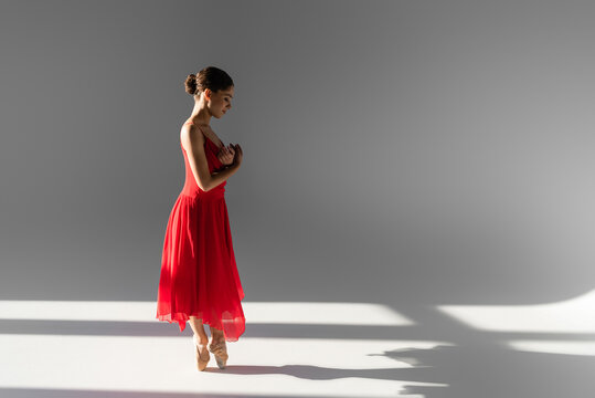 Side view of young ballerina dancing in red dress and pointe shoes on grey background with sunlight