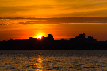 Fototapeta premium Amazing golden sunrise or sunset at the city. Black silhouette of the city on the sea shore. Colorful sky with clouds.