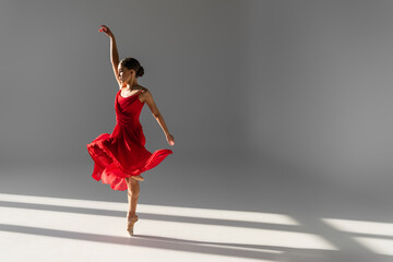 Side view of young ballerina in red dress dancing on grey background with sunlight