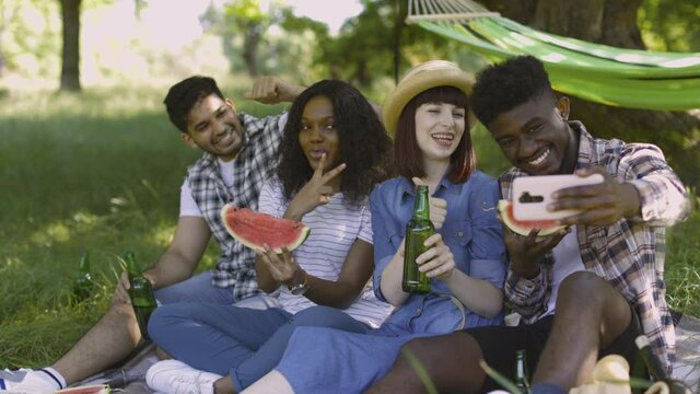 Four diverse friends sitting at green garden with watermelon and beer in hands and taking selfie on modern smartphone or having call with friend. Concept of people, technology and leisure time.