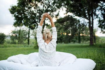 Young blonde woman wake up wear pajamas sitting in bed on white blanket outdoors over green nature...