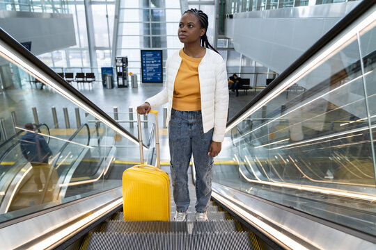 Millennial black woman with yellow suitcase on escalator in airport terminal or train station travel. Back to normal in tourism. Young afro female tourist before flight departure.