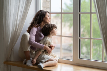 Curious mom and kid boy looking outside sitting on window sill. Caring mother or babysitter exploring world from inside on quarantine at home. Young mum and child son wait for dad return from work