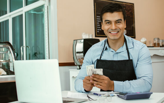 Caucasian handsome male bakery cafe business owner or entrepreneur wearing blue apron and eyeglasses, smiling with happiness and success, holding money in his hand, calculating profits while sitting. - Powered by Adobe