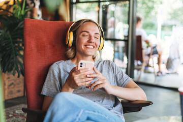 Young smiling blonde woman with close eyes in yellow headphones enjoys music with mobile phone...