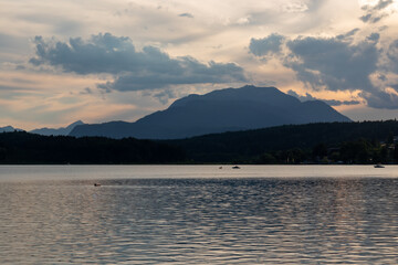 A panoramic view on the Lake Faak in Austria. The lake is surrounded by high Alpine peaks. The sun in slowly setting behind the mountains. Lots of clouds. Calm surface reflects the sunbeams. Serenity