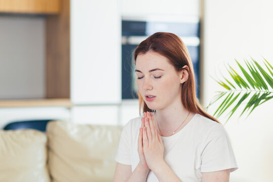 Closeup portrait of a young woman praying sincerely with folded arms at home or indoors. Sincere prayer of a Caucasian girl or female in the church with closed eyes alone. close up at home in the room