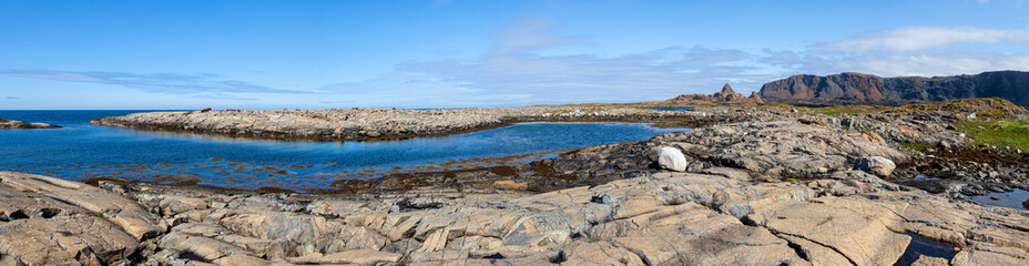 Fototapeta na wymiar Wide panorama. Rocky stone coast on the shore of a blue fjord with an island on the horizon under a blue sky. A mountain range with a lonely peak. Leka Island. Norway.