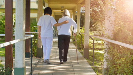 Asian doctor helping elderly retired woman with walker stick in the hospital yard. Rehabilitation concept